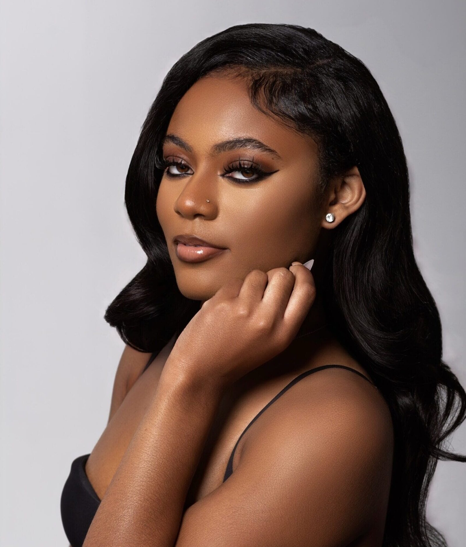 Portrait of Mya Riggins with long wavy hair, wearing a black strap top and resting her chin on her hand.